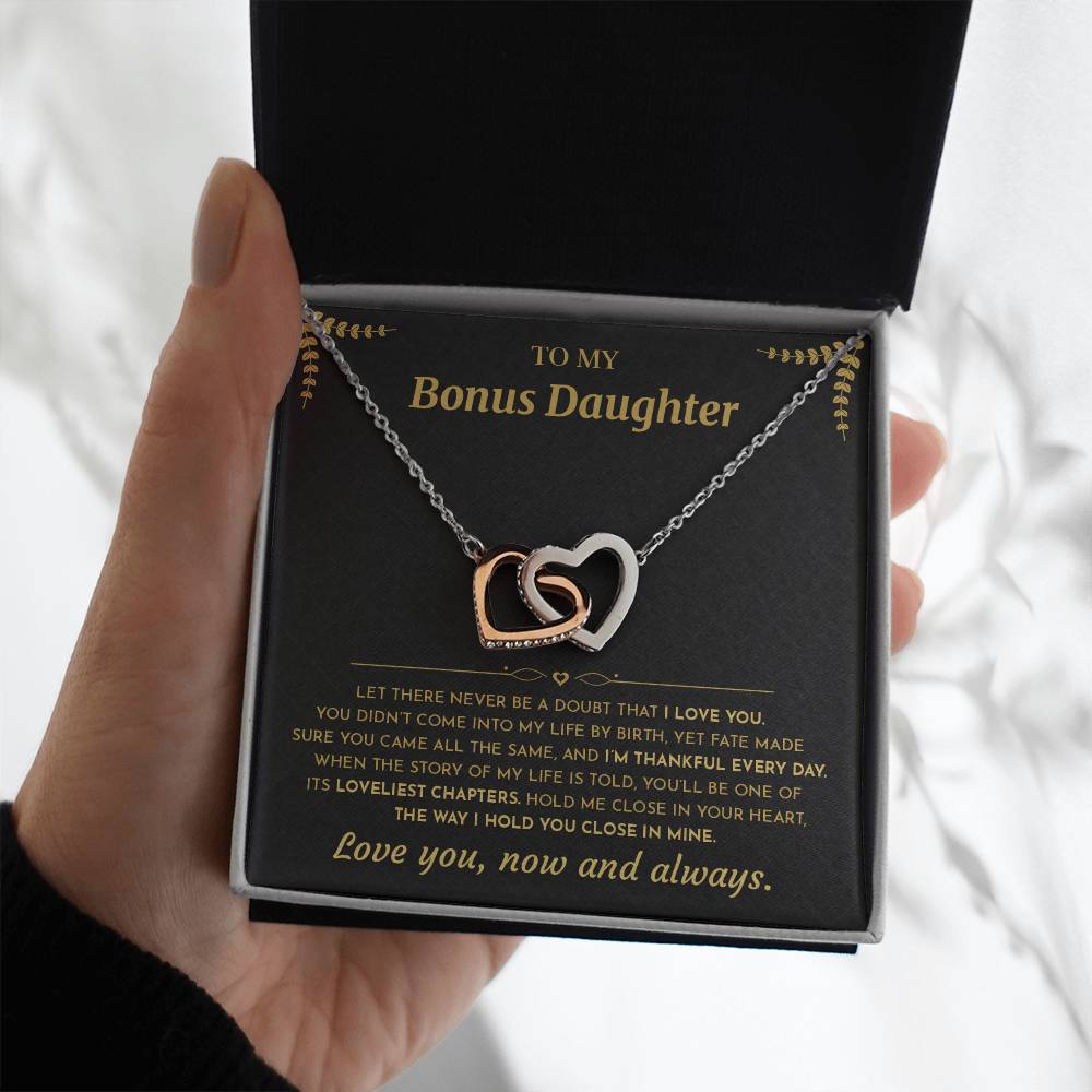 Hand holding the black and gold To My Bonus Daughter card with the Interlocking Hearts necklace in a black gift box