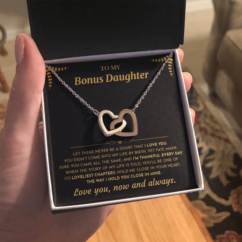Open gift box with the black and gold To My Bonus Daughter card and Interlocking Hearts necklace held up indoors