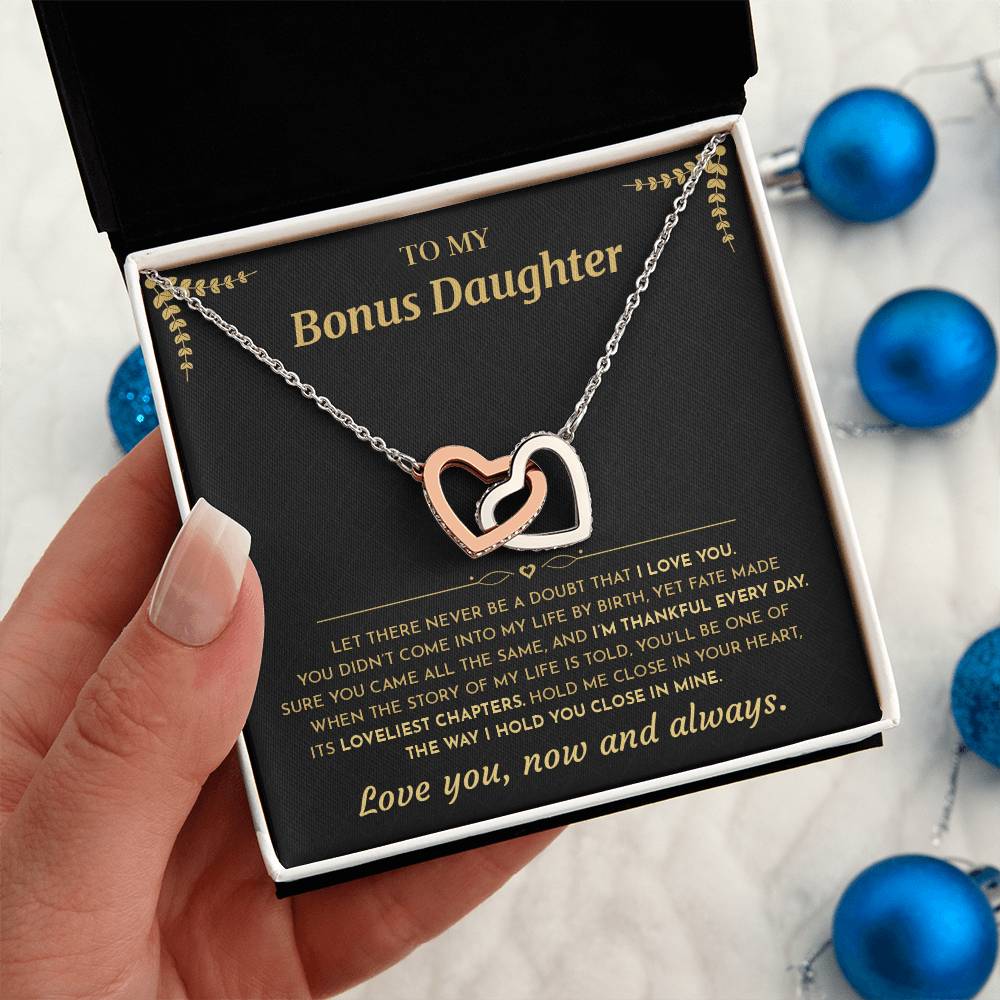 Hand holding the black and gold To My Bonus Daughter card gift box beside blue Christmas ornaments