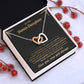 Tilted white gift box holding the black and gold To My Bonus Daughter card and Interlocking Hearts necklace among red berries and pinecones