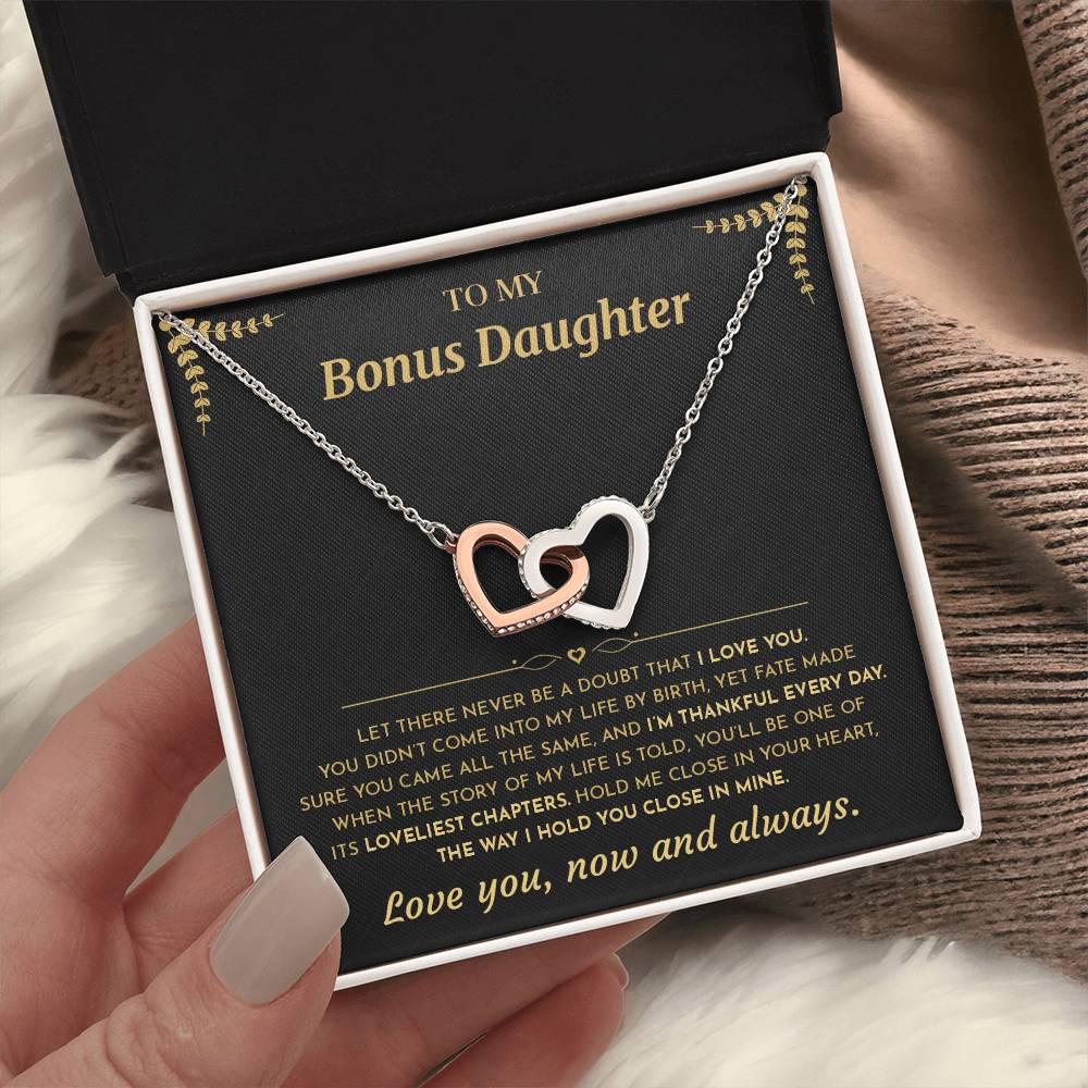 Hand holding an open black gift box with the black and gold To My Bonus Daughter card and linked hearts necklace beside a knit blanket