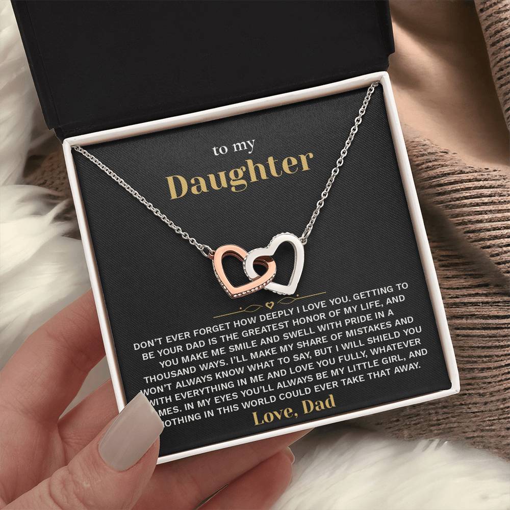 Hand holding an open black gift box with the black and gold To My Daughter card from Dad and linked hearts necklace beside a knit blanket