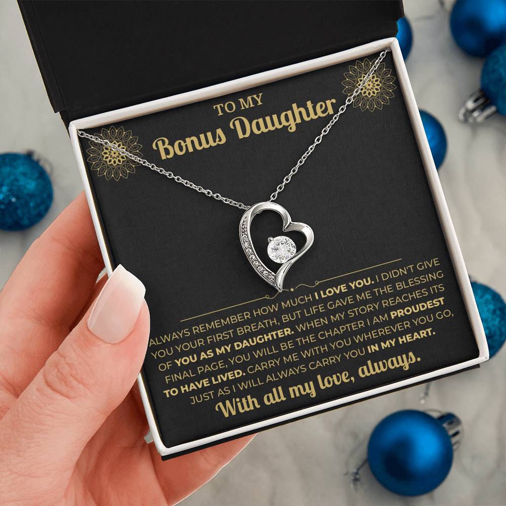 Hand holding the black and gold To My Bonus Daughter card gift box by blue Christmas ornaments on white fur