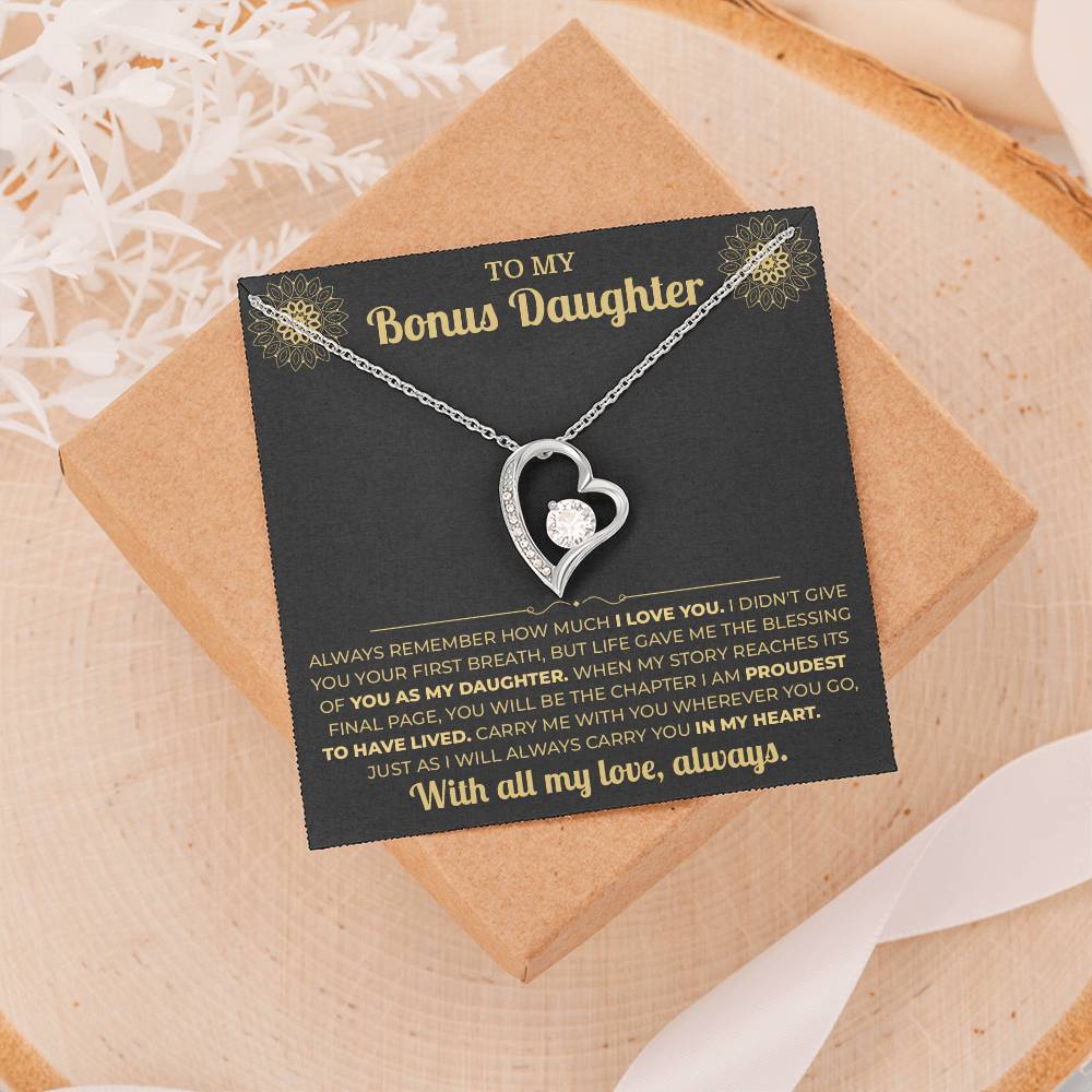 Black and gold To My Bonus Daughter card and heart necklace on a gift box resting on a wood slice with dried flowers