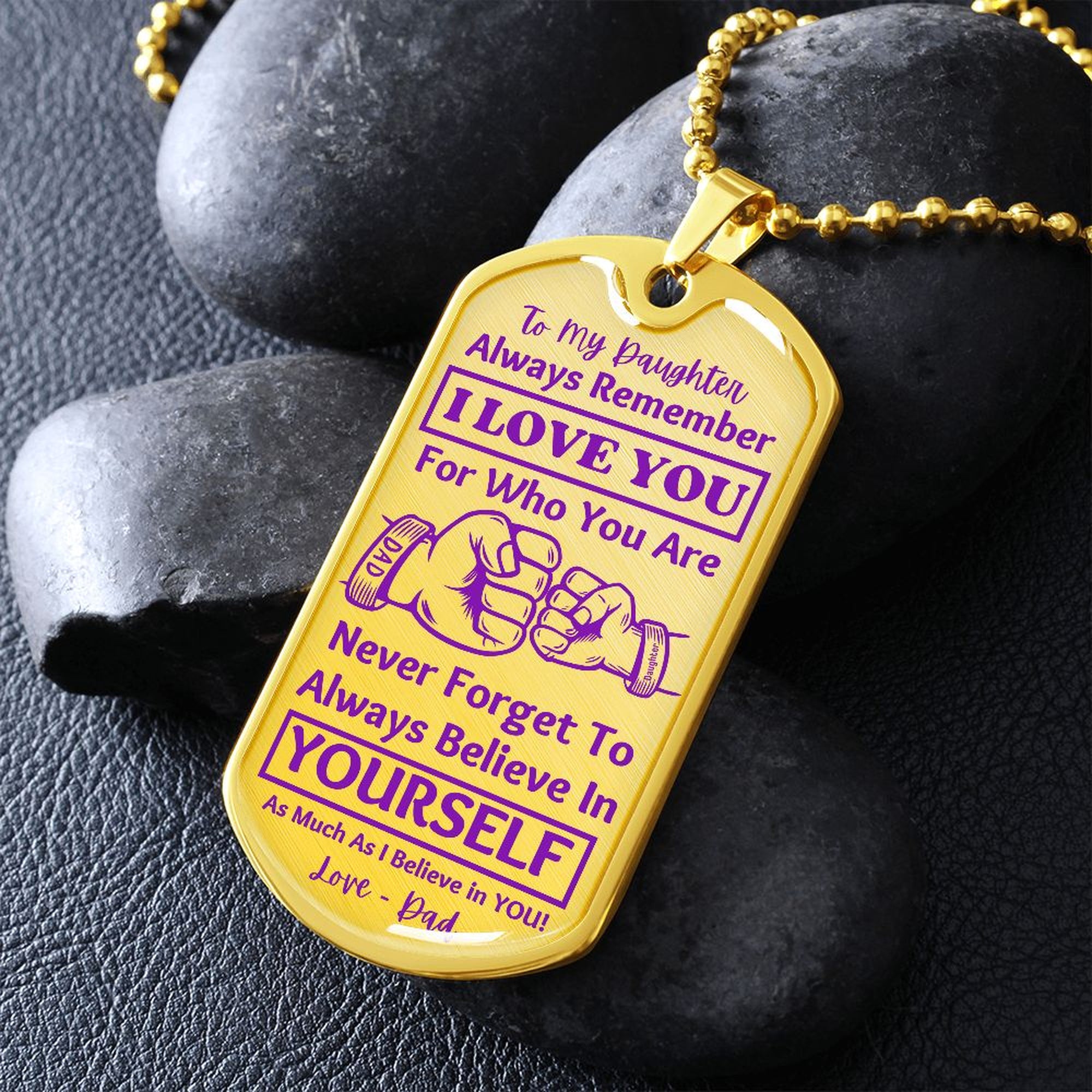 Gold-finish I Love You Daughter dog tag propped on dark river rocks